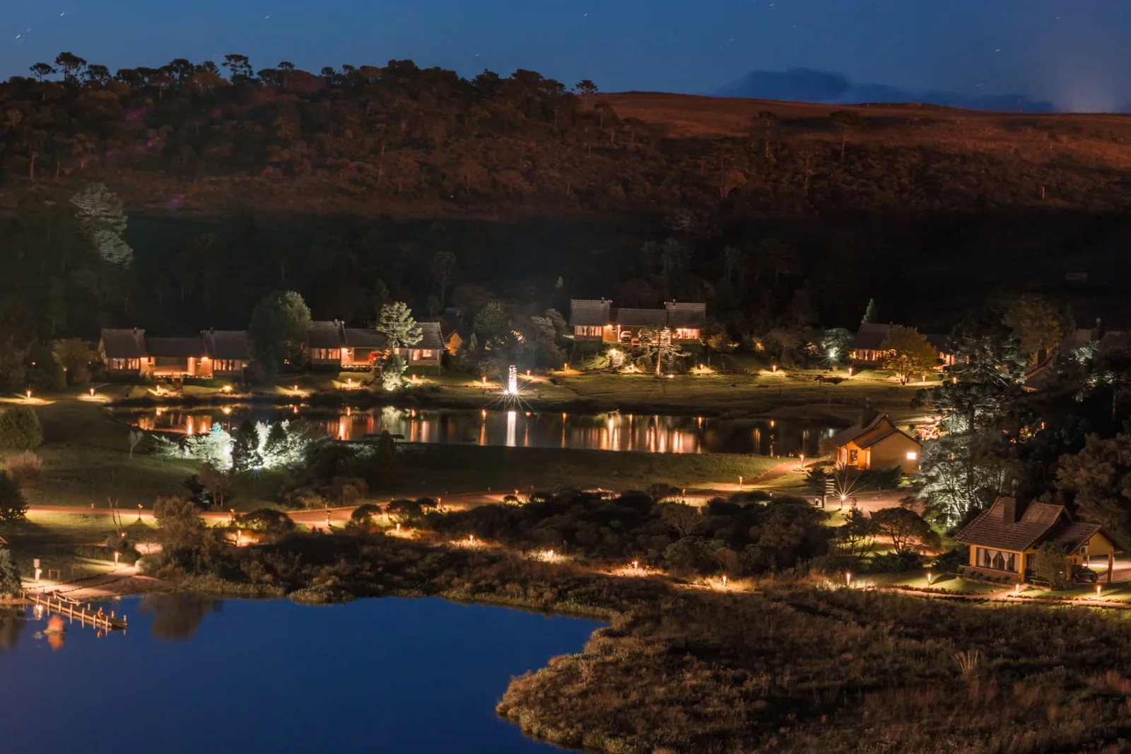 Vista noturna do Rio do Rastro Eco Resort iluminado entre o lago e a paisagem da Serra Catarinense